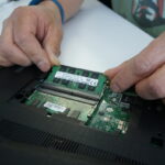 A close-up photo of the inside of a laptop. A hand is shown carefully unplugging the battery connector from the motherboard to ensure safety during the RAM upgrade