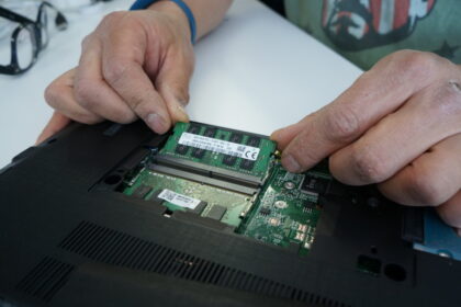 A close-up photo of the inside of a laptop. A hand is shown carefully unplugging the battery connector from the motherboard to ensure safety during the RAM upgrade