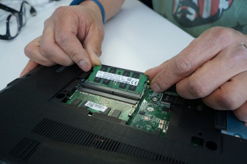 A close-up photo of the inside of a laptop. A hand is shown carefully unplugging the battery connector from the motherboard to ensure safety during the RAM upgrade