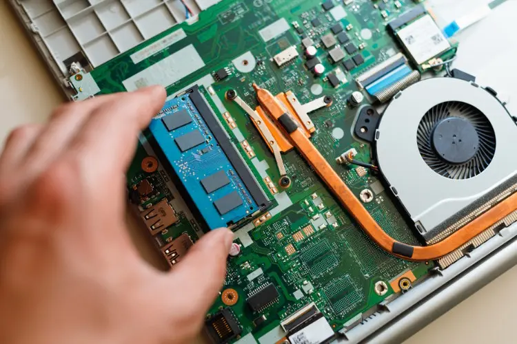 A close-up of a green SO-DIMM RAM module being carefully placed into a slot on an open laptop motherboard, illustrating the process of a RAM upgrade.